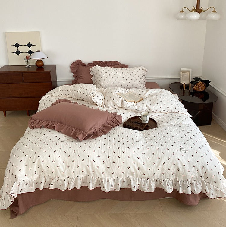A bed with a white quilt cover featuring a red cherry pattern, accompanied by brown and white pillows, in a room with a wooden floor and a brown bedside table.