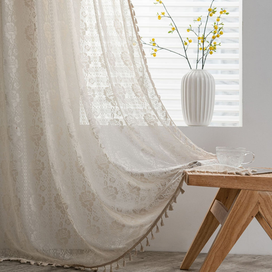 Lace curtain with tassels in a room with a wooden table and vase of flowers.