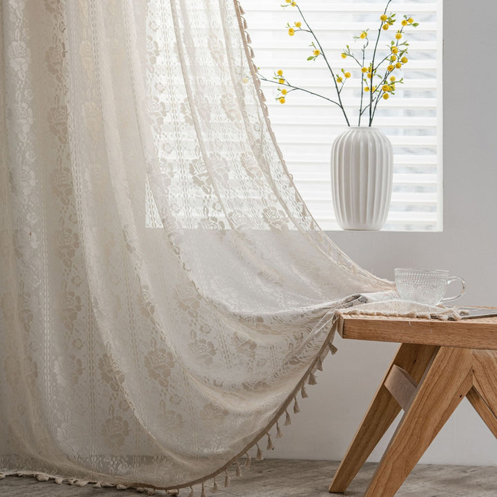 Lace curtain with tassels in a room with a wooden table and vase of flowers.