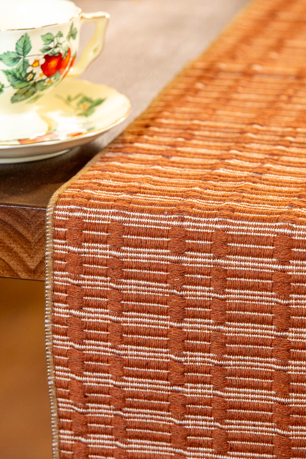 A close-up of a woven table runner in a warm rust-orange and cream pattern, draped over a wooden table next to a floral teacup and saucer.