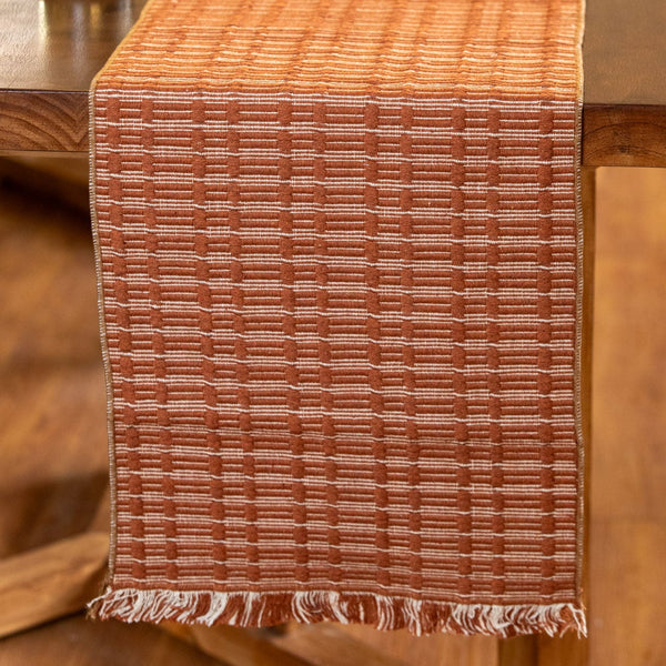 A woven rectangular table runner in a warm rust-orange and beige pattern, draped over a dark wooden dining table with a small plant in the background.
