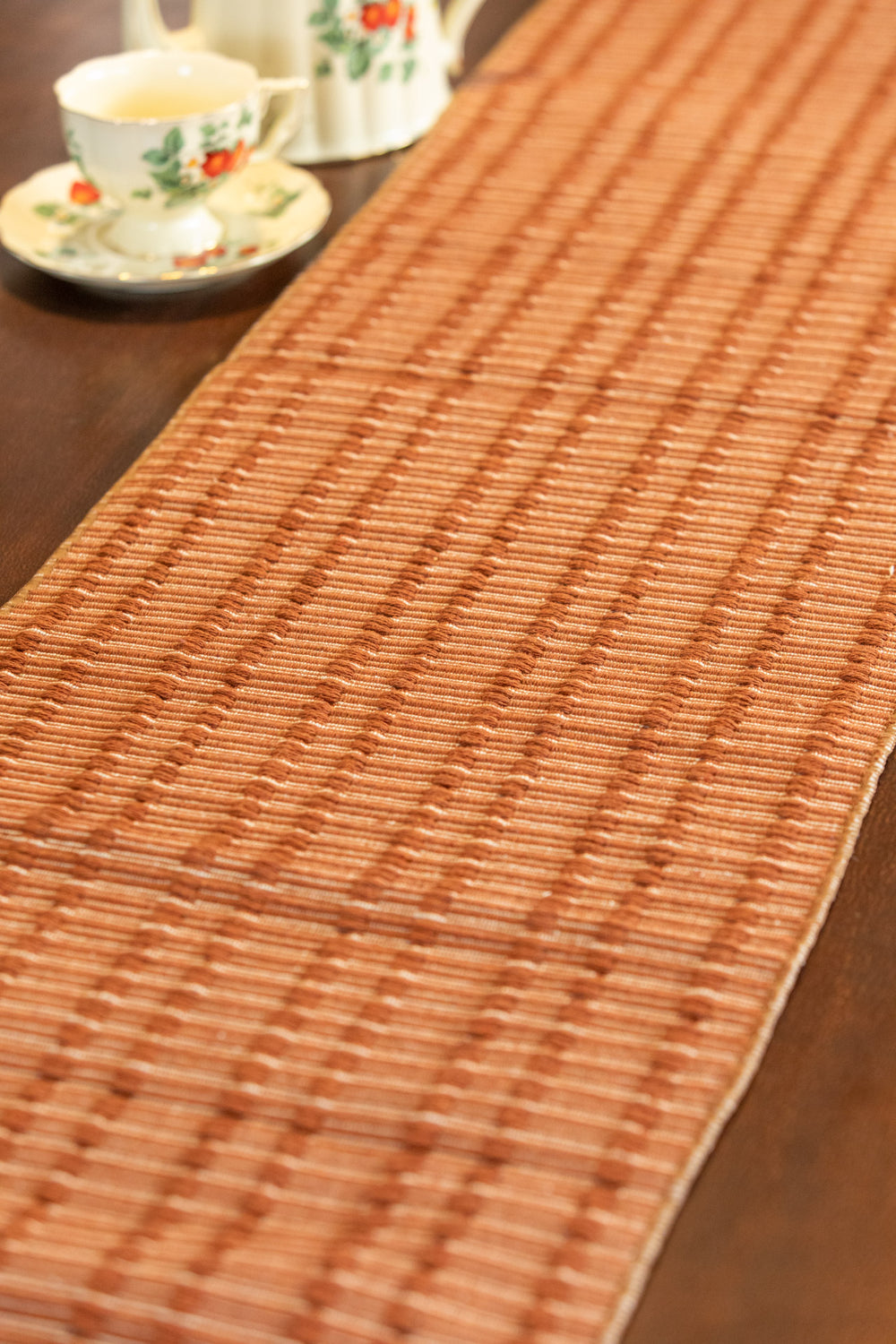 A woven brown table runner with a textured basket-weave pattern, laid diagonally on a dark wooden table with a teacup and saucer in the background.