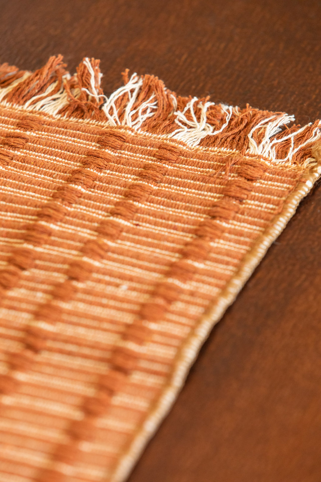 Close-up of a woven textile with fringed edges in warm tan and cream tones, laid on a dark brown wooden surface.