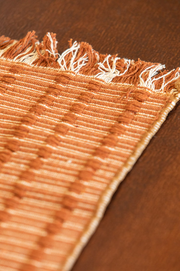 Close-up of a woven textile with fringed edges in warm tan and cream tones, laid on a dark brown wooden surface.