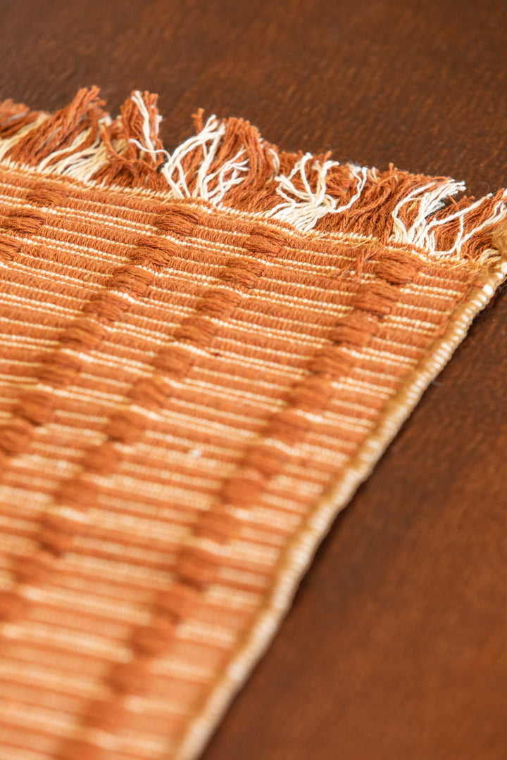 Close-up of a woven textile with fringed edges in warm tan and cream tones, laid on a dark brown wooden surface.