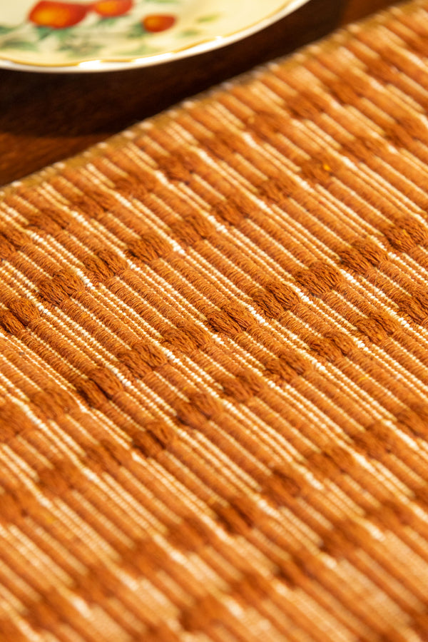 A close-up of a woven bamboo placemat in warm tan and brown tones, with a small portion of a decorative plate visible in the upper corner.