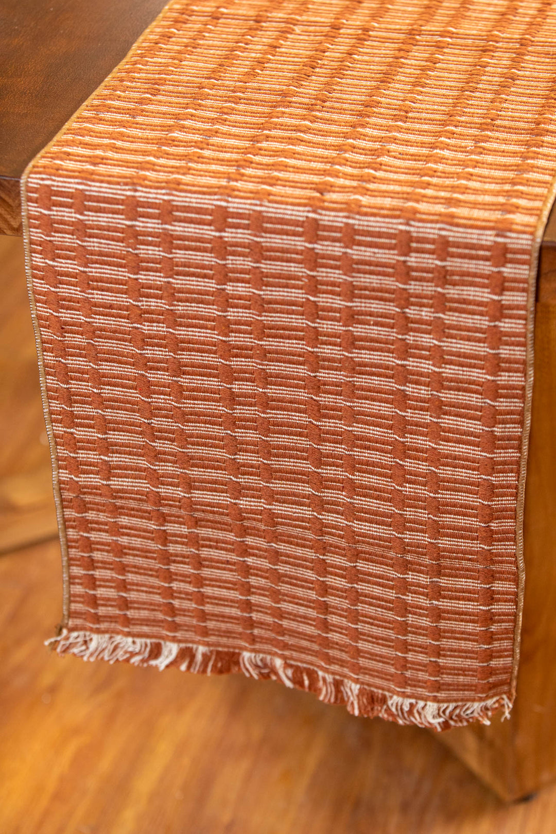 A close-up of a woven table runner in a warm rust-orange and beige pattern, draped over a wooden table with a matching wooden floor visible in the background.