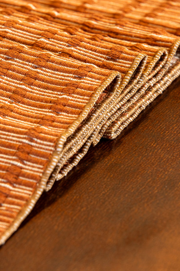 A close-up of a folded woven placemat or rug in warm brown and tan tones, showing the ribbed texture and layered edges on a dark brown surface.