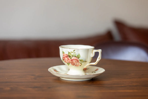 A vintage-style ceramic teacup and saucer set with a pink rose floral pattern, resting on a wooden table with a blurred background.