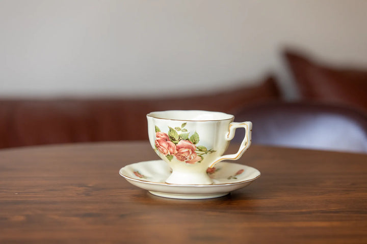 A vintage-style ceramic teacup and saucer set with a pink rose floral pattern, resting on a wooden table with a blurred background.