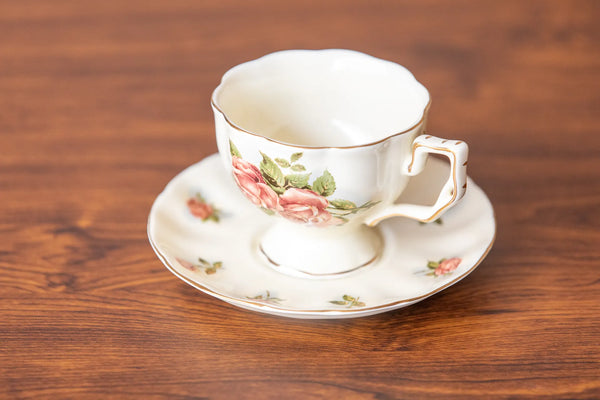 A vintage-style white porcelain teacup and saucer set with a pink rose floral pattern and gold trim, displayed on a rustic wooden surface.